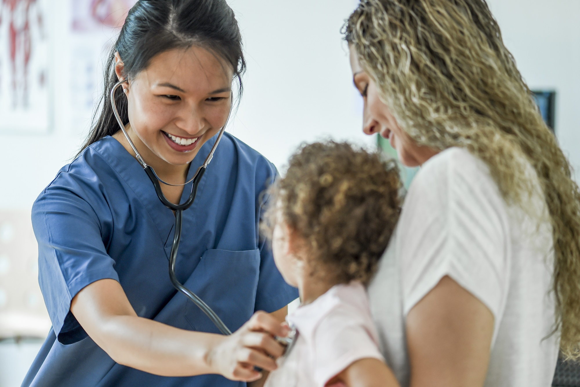 A little girl is with her mom at a check up appointment at the doctors office. A nurse is listening to the little girls heartbeat.